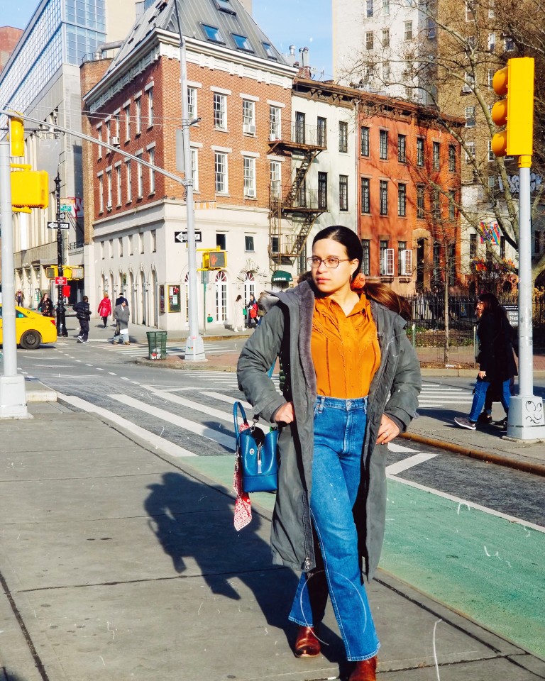 Brunette strolls sidewalk in gray parka, dark blue jeans, and coral blouse.